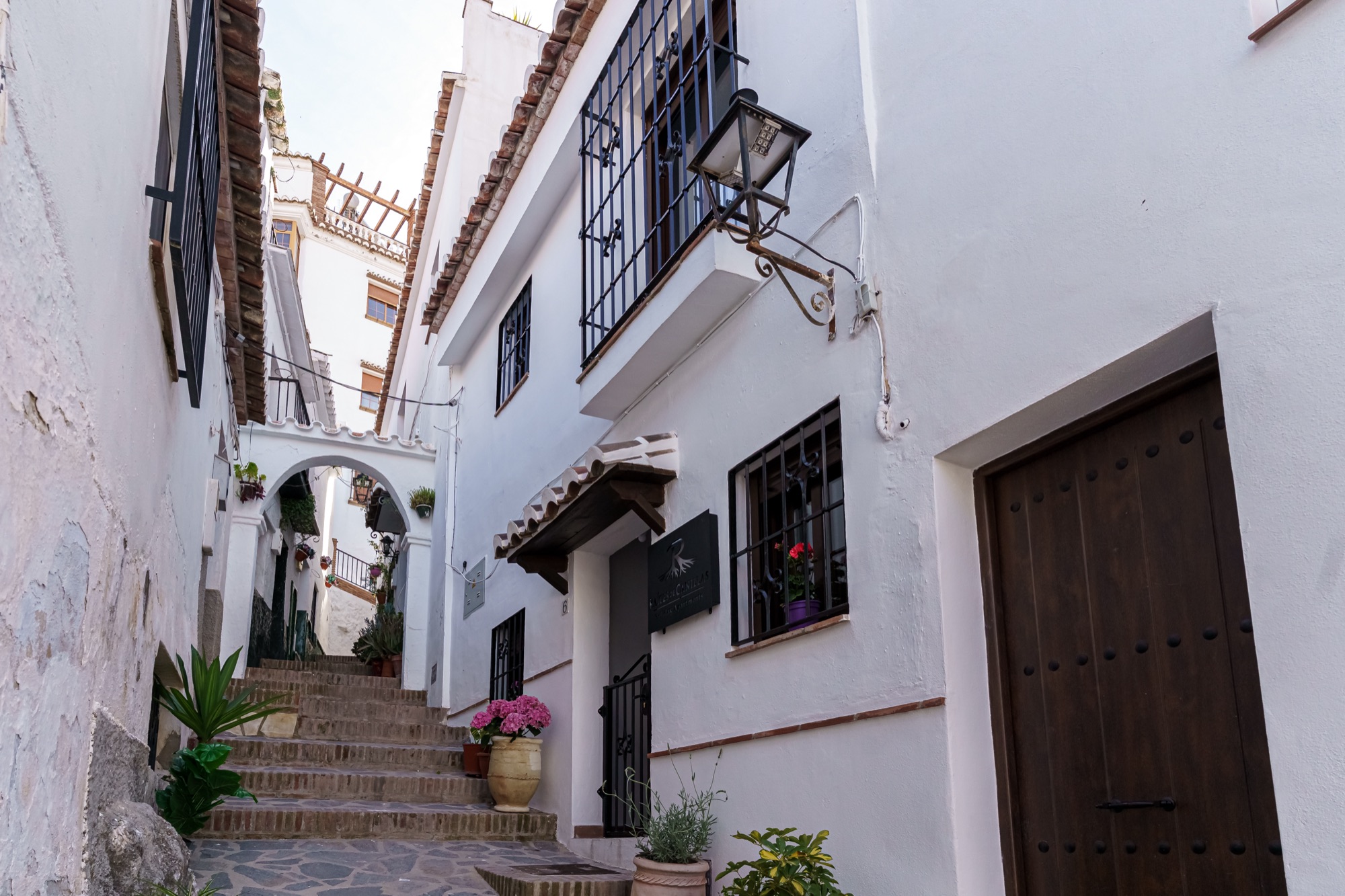 Calleja empedrada y en escalera del pueblo, con casas encaladas, rejas negras, farol de forja, arco al fondo y la entrada del alojamiento con su tejadillo y macetas floridas.