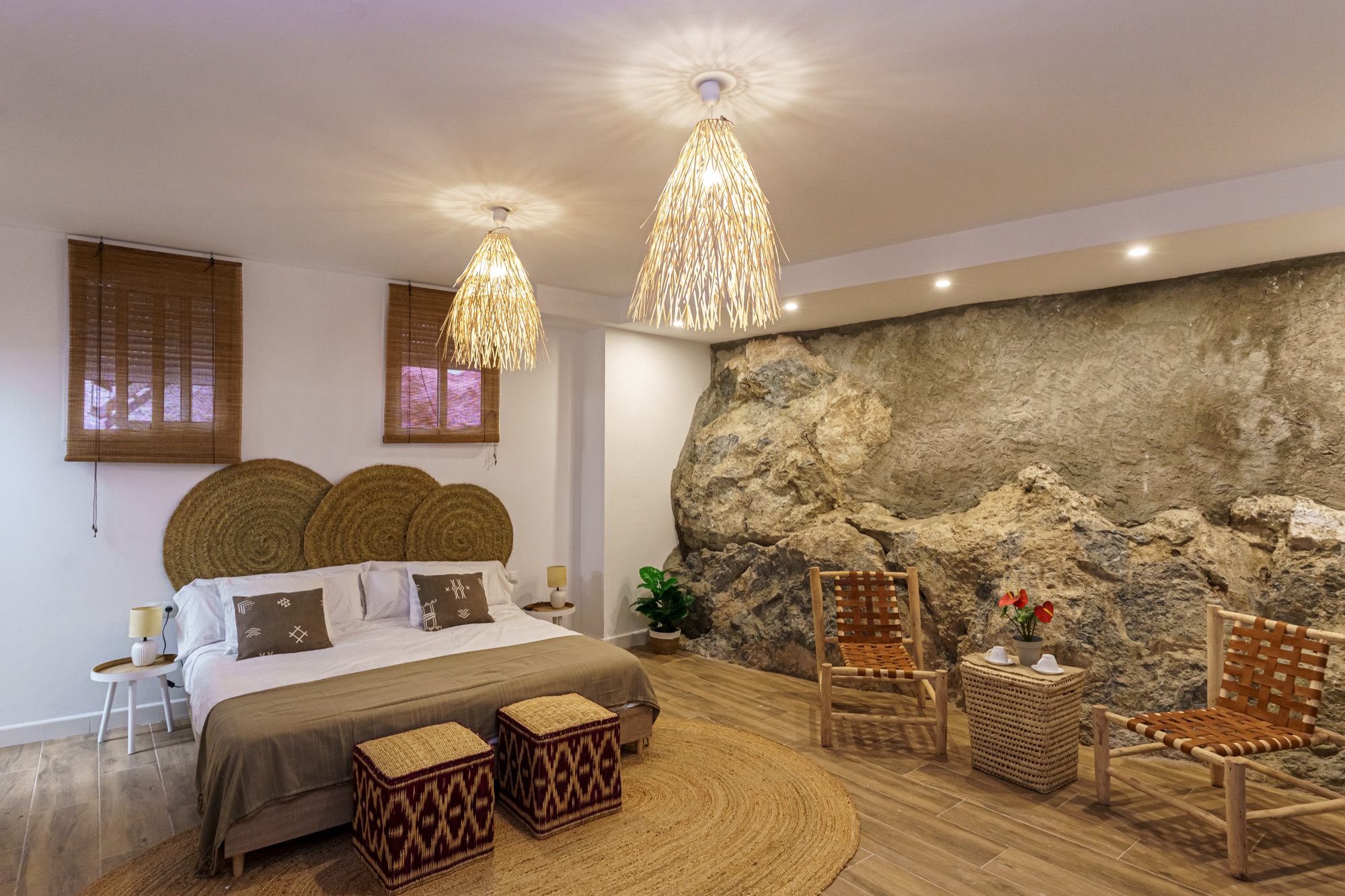 Suite Maroma: cama con triple cabecero de esparto, dos lámparas colgantes de fibra natural encendidas y la imponente pared de roca natural vista con dos sillas artesanales.