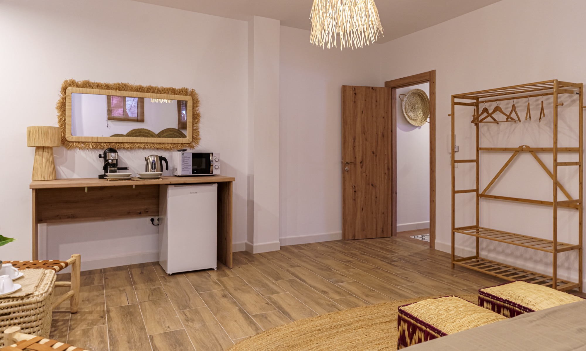 Zona de office del apartamento con escritorio de madera, cafetera, hervidor, microondas y minibar, espejo con marco de rafia y burro de bambú junto a la puerta de entrada.