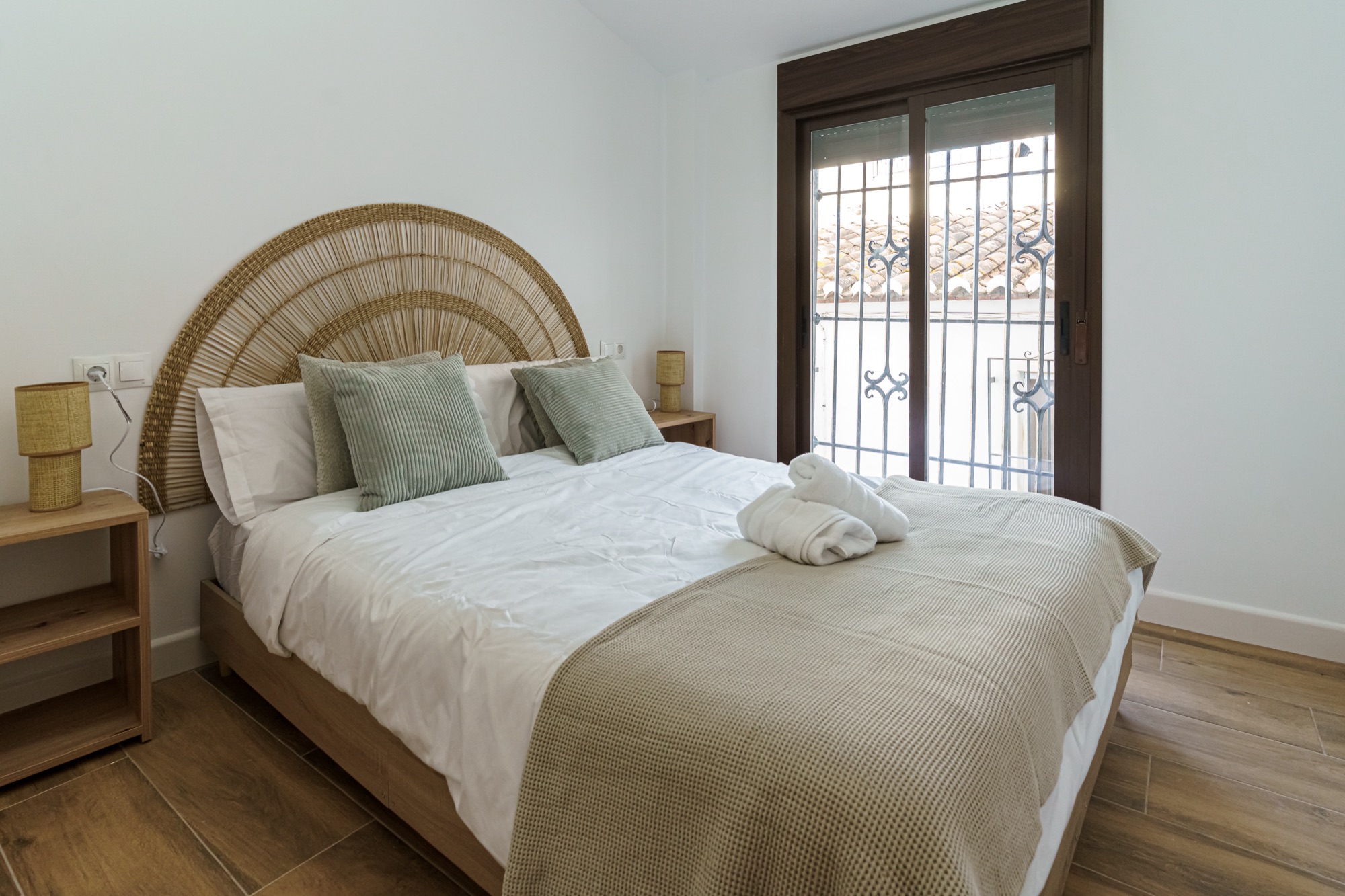 Dormitorio del apartamento Olivo con cama doble, espectacular cabecero artesanal de fibra en forma de abanico, cojines verde salvia, toallas enrolladas sobre la colcha y ventanal con reja andaluza que asoma a los tejados del pueblo.