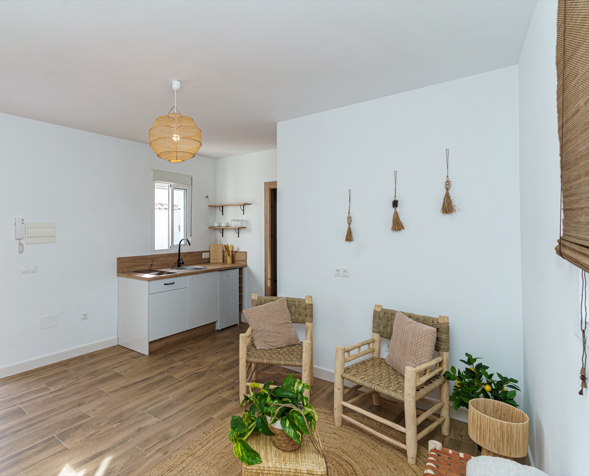 Vista general del salón con kitchenette de madera y frentes blancos, butacas de enea con cojines, plantas y escobones artesanales colgados en la pared blanca.