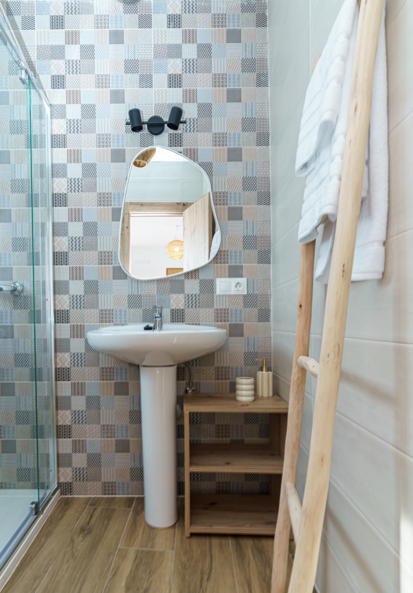Baño con pared de azulejo tipo patchwork en azules y grises, lavabo de pedestal blanco, espejo de forma orgánica, mampara de ducha y escalera de madera como toallero.