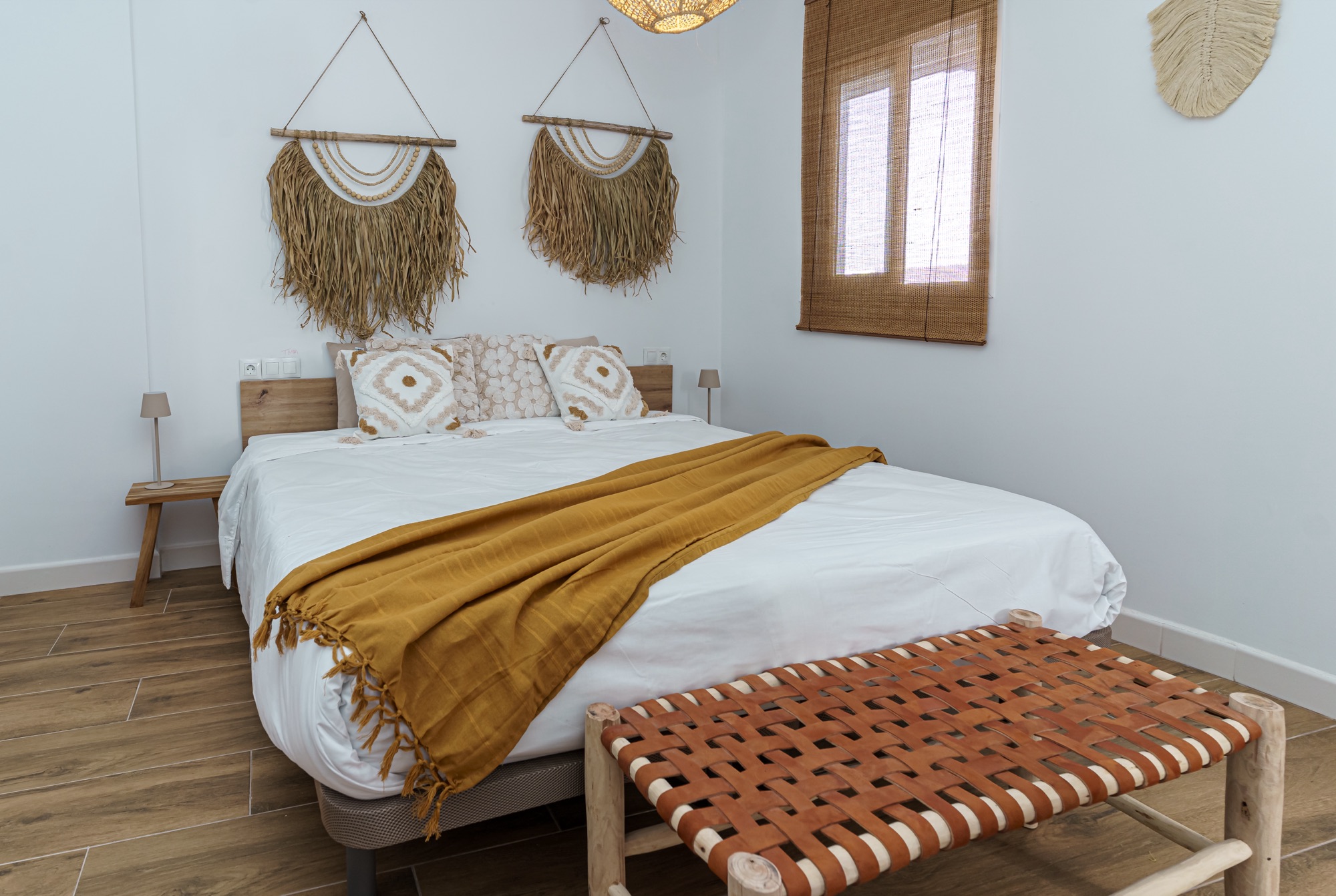 Dormitorio en primer plano: cama con cojines boho, manta mostaza con flecos, tapices de rafia colgados sobre el cabecero de madera y banqueta artesanal de cuero trenzado a los pies.