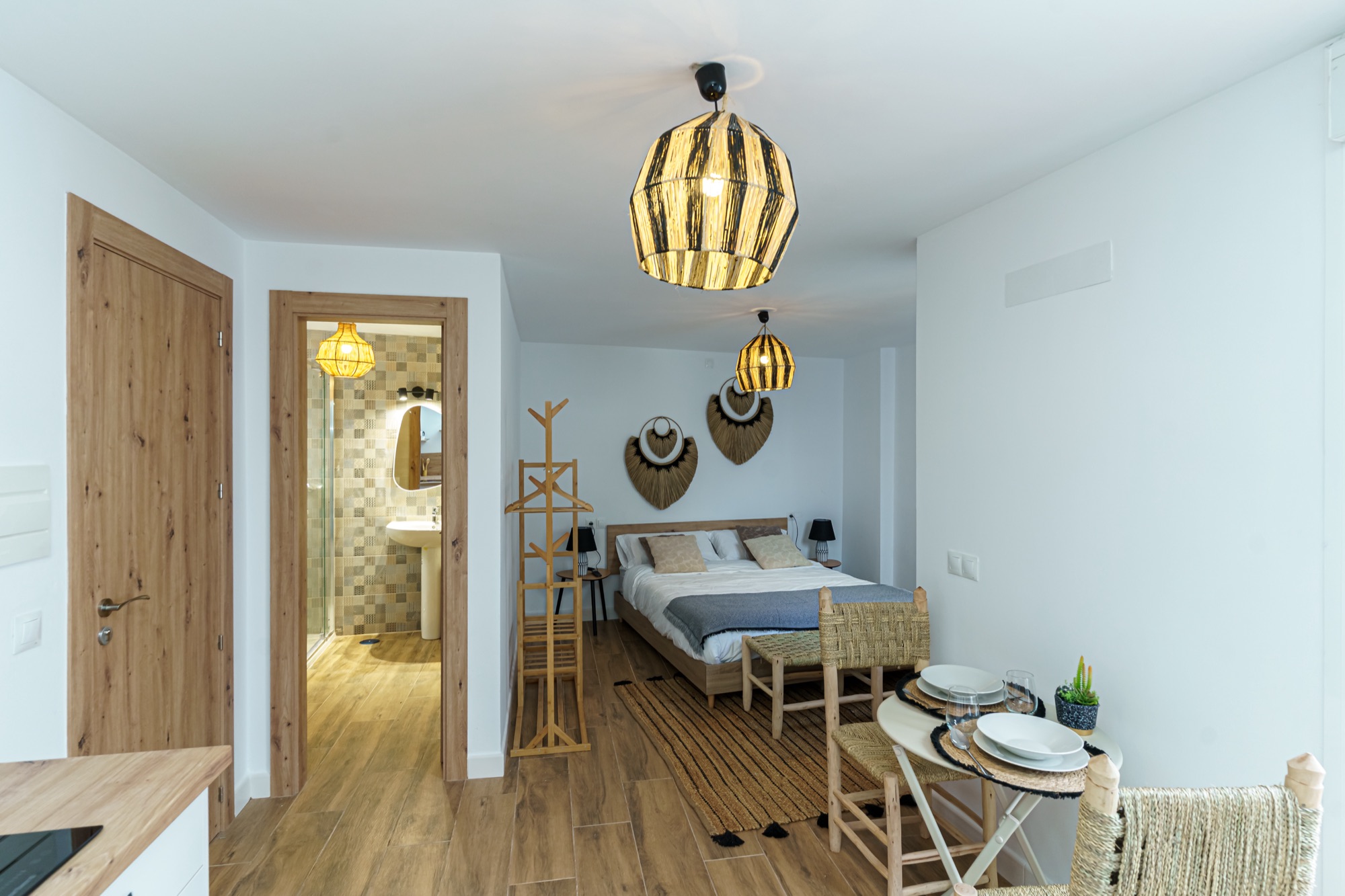 Vista general del estudio Saltillo: cama doble con plaid gris, mesita de comedor montada con vajilla, lámparas colgantes de fibra natural y baño al fondo, todo en estilo boho con madera clara.