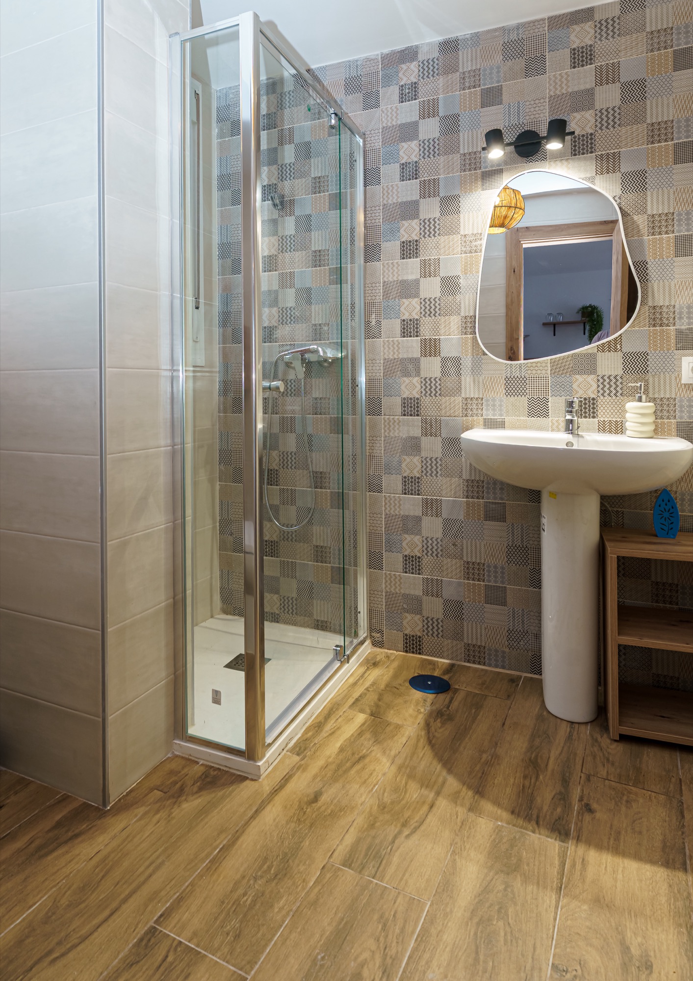 Baño con ducha de mampara transparente, lavabo de pedestal, espejo de forma orgánica con marco de madera y pared de azulejos tipo patchwork en tonos tierra sobre suelo imitación madera.