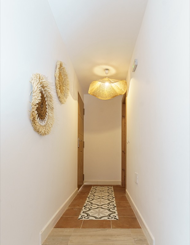 Pasillo distribuidor blanco con dos juju hats de plumas y fibras naturales en la pared, lámpara de ratán encendida, puertas de madera y alfombra de baldosa hidráulica embutida en el suelo de barro.