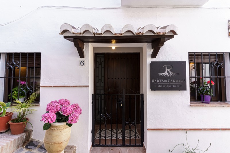 Fachada encalada de la entrada de Raíces de Canillas: puerta de madera oscura bajo tejadillo de teja árabe, cancela de forja, rótulo del alojamiento, rejas con geranios y una hortensia rosa en tinaja de barro.
