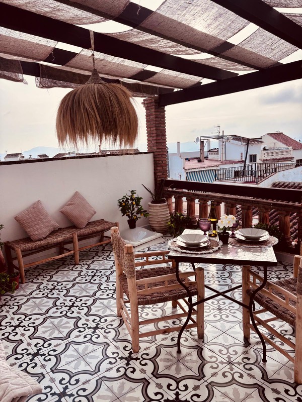 Terraza-azotea con pérgola de vigas oscuras y toldos de arpillera, mesa de forja vestida para dos con sillas marroquíes de madera y cuerda, suelo de baldosa hidráulica en blanco y negro y tejados del pueblo con montañas al fondo.