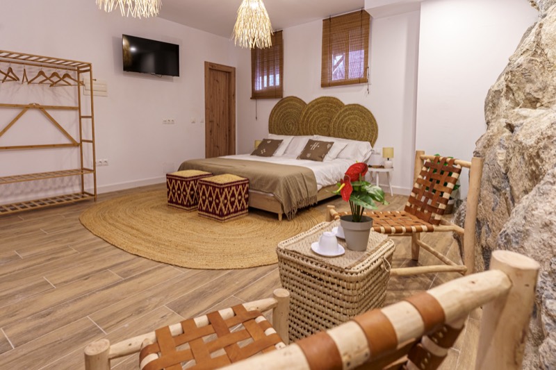 Dormitorio de Maroma visto desde el rincón de estar: sillas de madera y tiras de cuero en primer plano, mesita-cesta de mimbre con planta roja, cama al fondo y la pared de roca a la derecha.