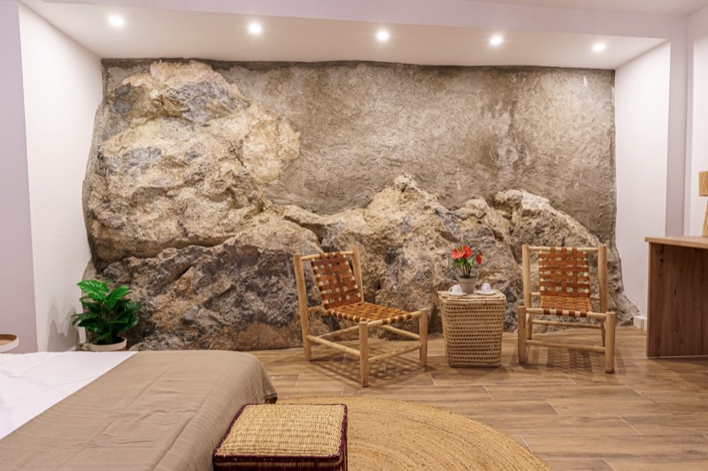La espectacular pared de roca natural integrada en la habitación, con dos sillas artesanales de cuero trenzado, mesita de fibra con flores y una planta, junto al borde de la cama.