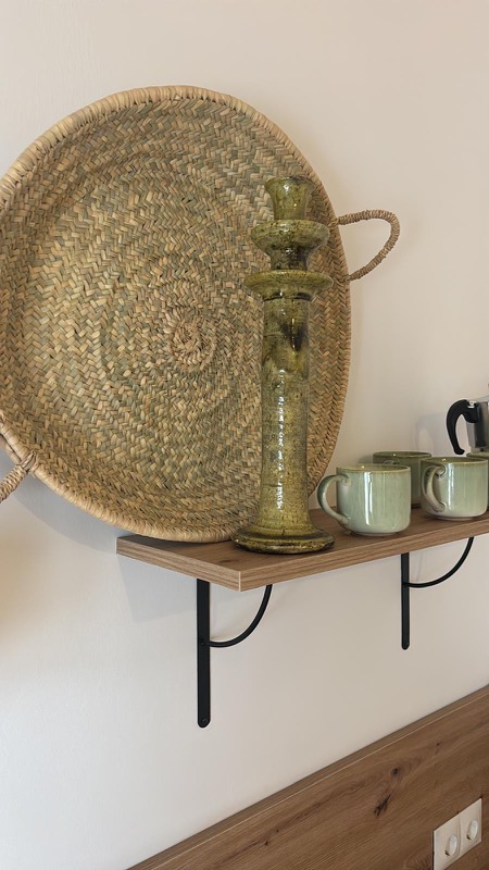 Detalle decorativo de una balda de madera con una gran bandeja de esparto trenzado colgada en la pared, un candelabro de cerámica verde oliva y tazas artesanales en tonos salvia junto a una cafetera italiana.