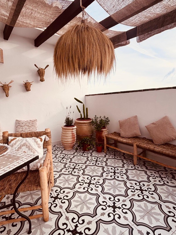 Terraza despejada con suelo hidráulico protagonista, bancos de madera y enea con cojines, macetas de barro con cactus y lavanda, y lámpara de rafia colgando de la pérgola.