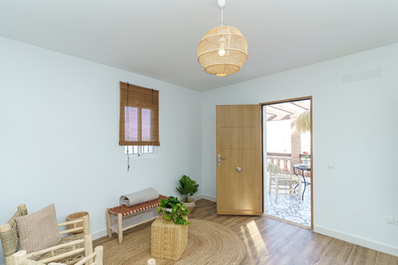 Salón con puerta de roble abierta hacia la terraza soleada, lámpara colgante de ratán, banco con esterilla enrollada y plantas sobre alfombra de yute.