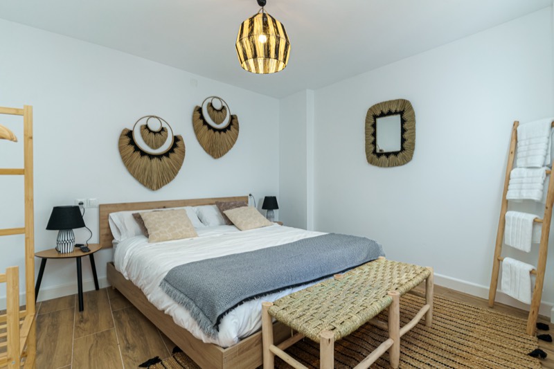 Dormitorio con cama doble vestida en blanco y gris, cabecero de madera, abanicos decorativos de fibra en la pared, banco de enea a los pies y escalera de madera con toallas.