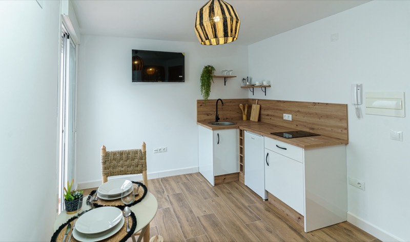 Rincón de cocina americana con encimera de madera, placa de inducción, minibar, fregadero con baldas abiertas, televisor en pared y mesa redonda con dos servicios junto a la ventana.